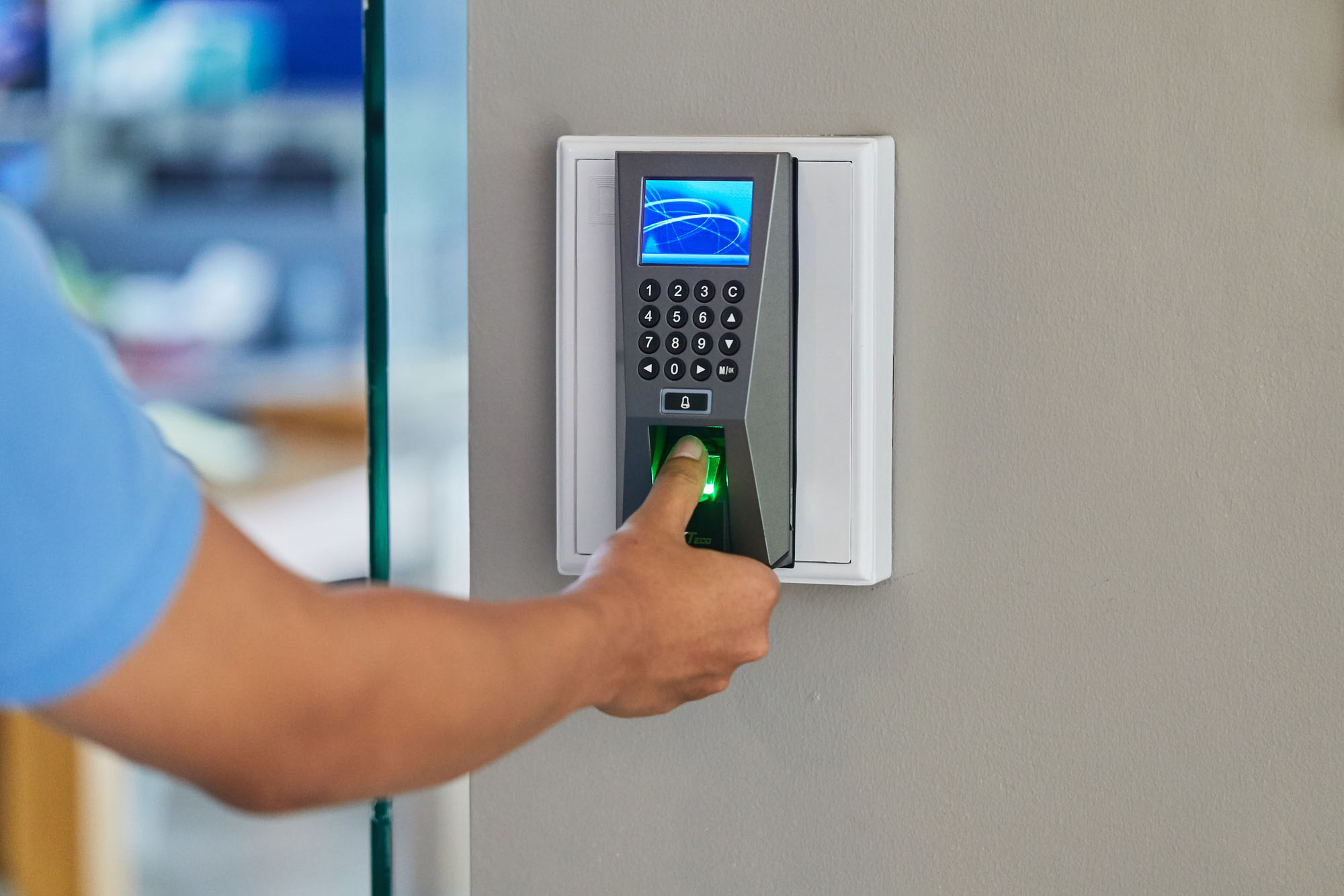 A person uses a biometric fingerprint scanner, pressing their finger against the green sensor to gain access, indicating secure entry.