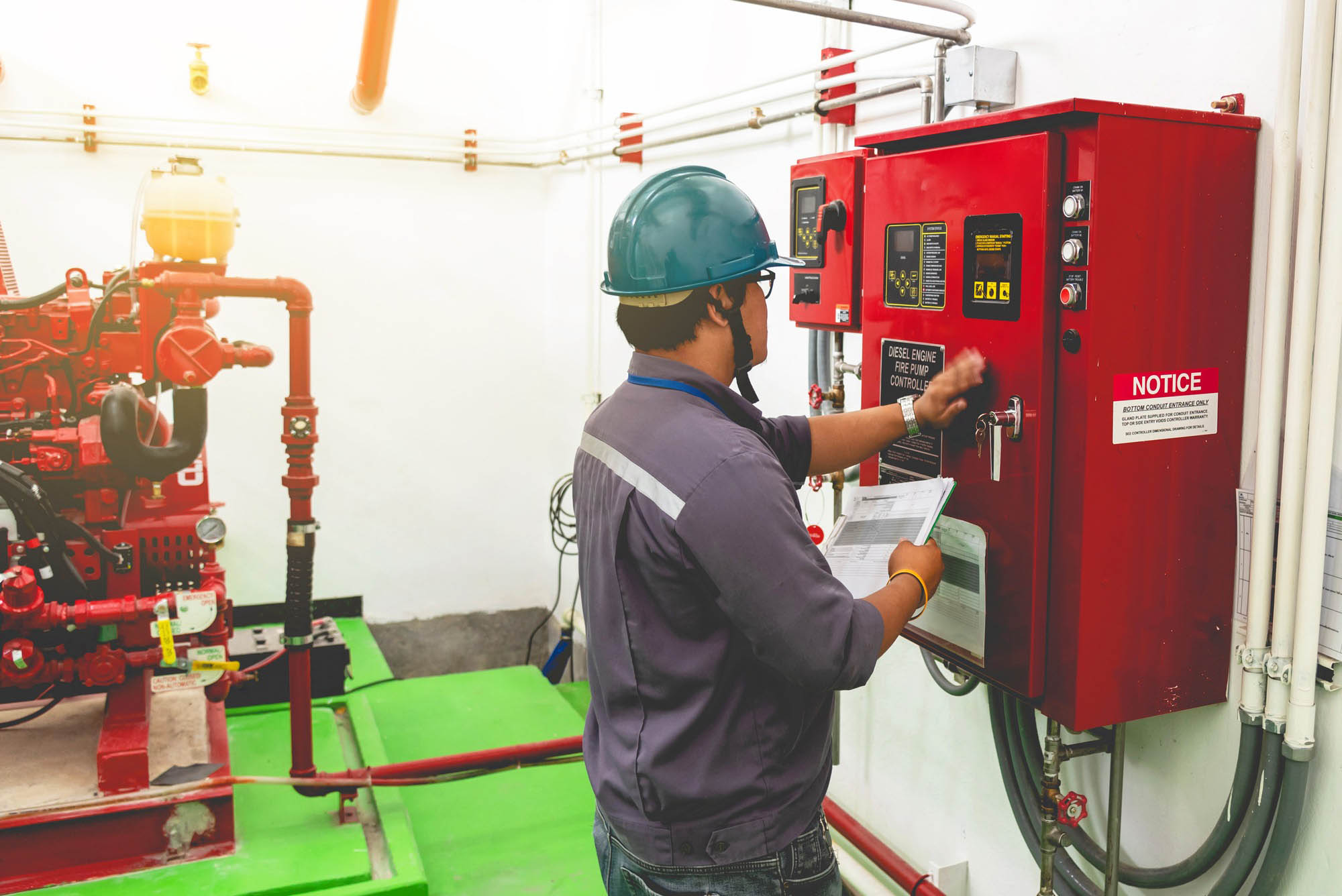 A technician in a hard hat and work attire operates a diesel engine fire pump control panel, reviewing documents to ensure proper functionality.